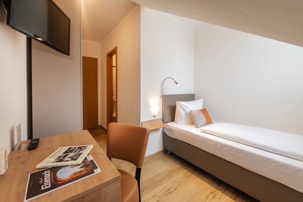 A small, modern hotel room with a single bed, a desk with a chair, a flat-screen TV mounted on the wall, and a bedside table with a lamp.