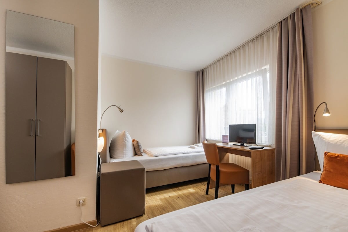 A hotel room with a bed, a desk with a computer, a chair, a wardrobe, and a window with curtains.
