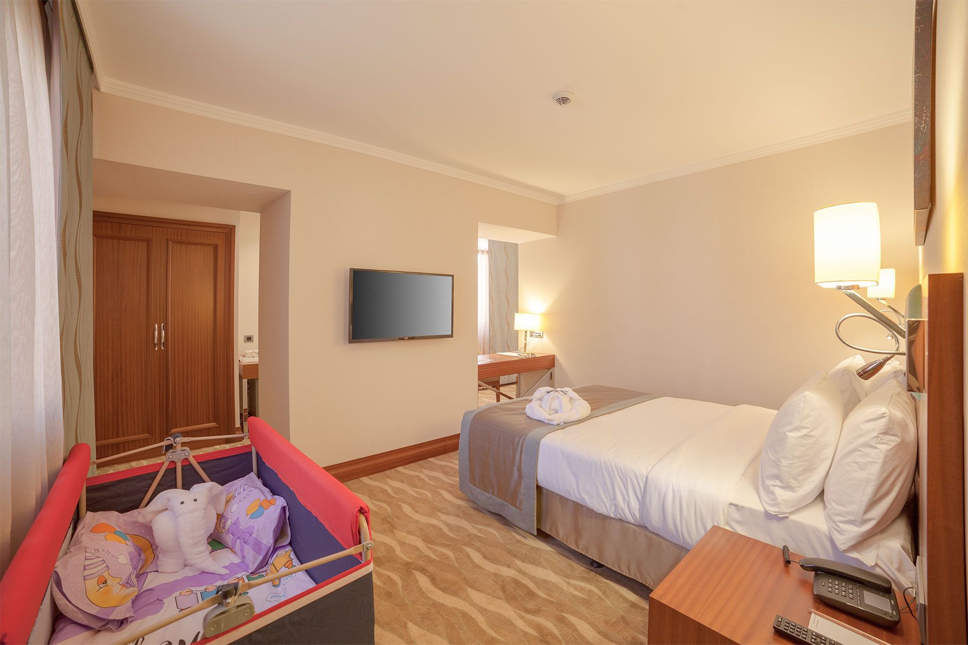A hotel room with a bed, a desk with a lamp, a television on the wall, and a wardrobe.