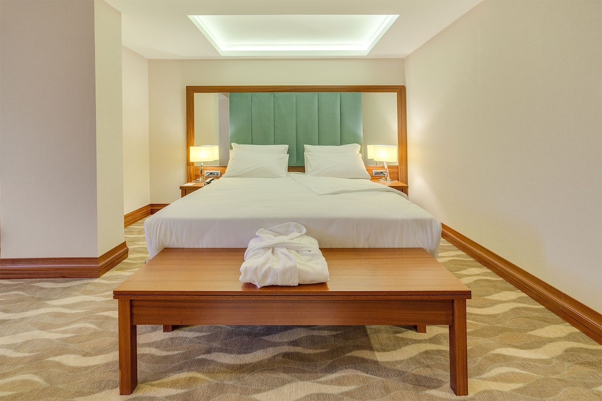 A hotel room with a large bed, two bedside tables with lamps, a wooden nightstand, and a small table with a towel.