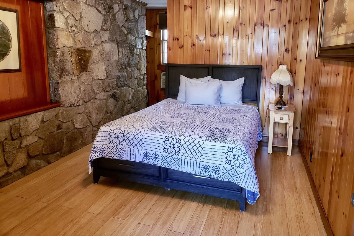 Ein Schlafzimmer mit Steinwand, Holzvertäfelung, einem Bett mit gemustertem Überwurf, einem Nachttisch mit Lampe und einem gerahmten Bild an der Wand.