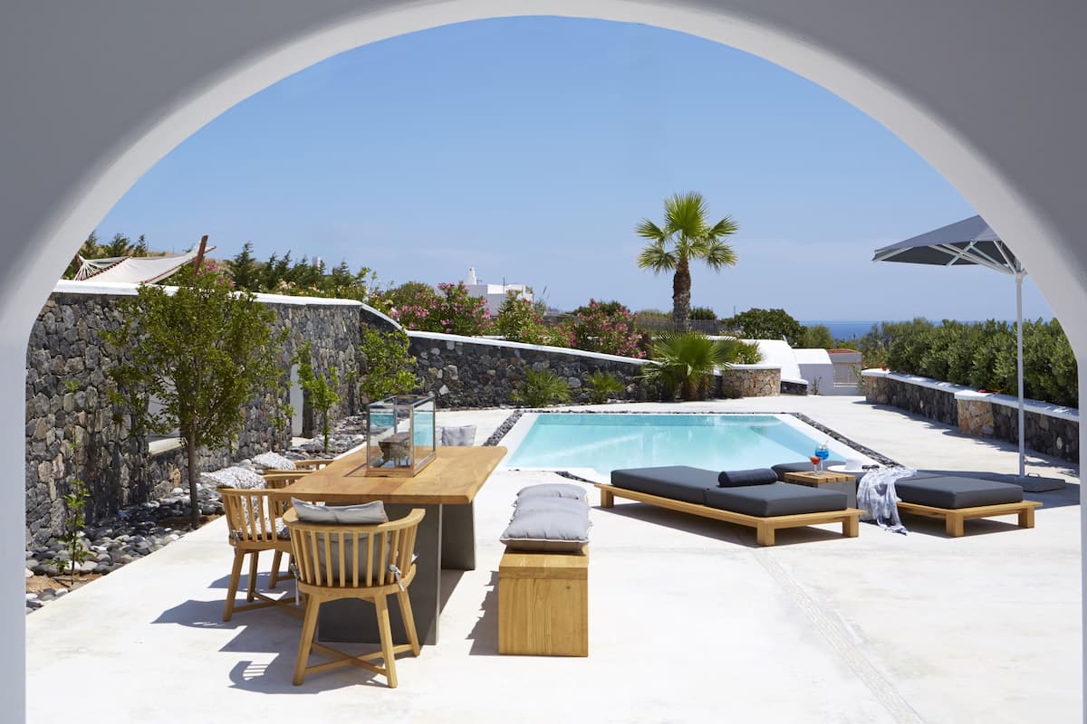 A poolside area with a stone wall, wooden furniture, and a view of the sea.