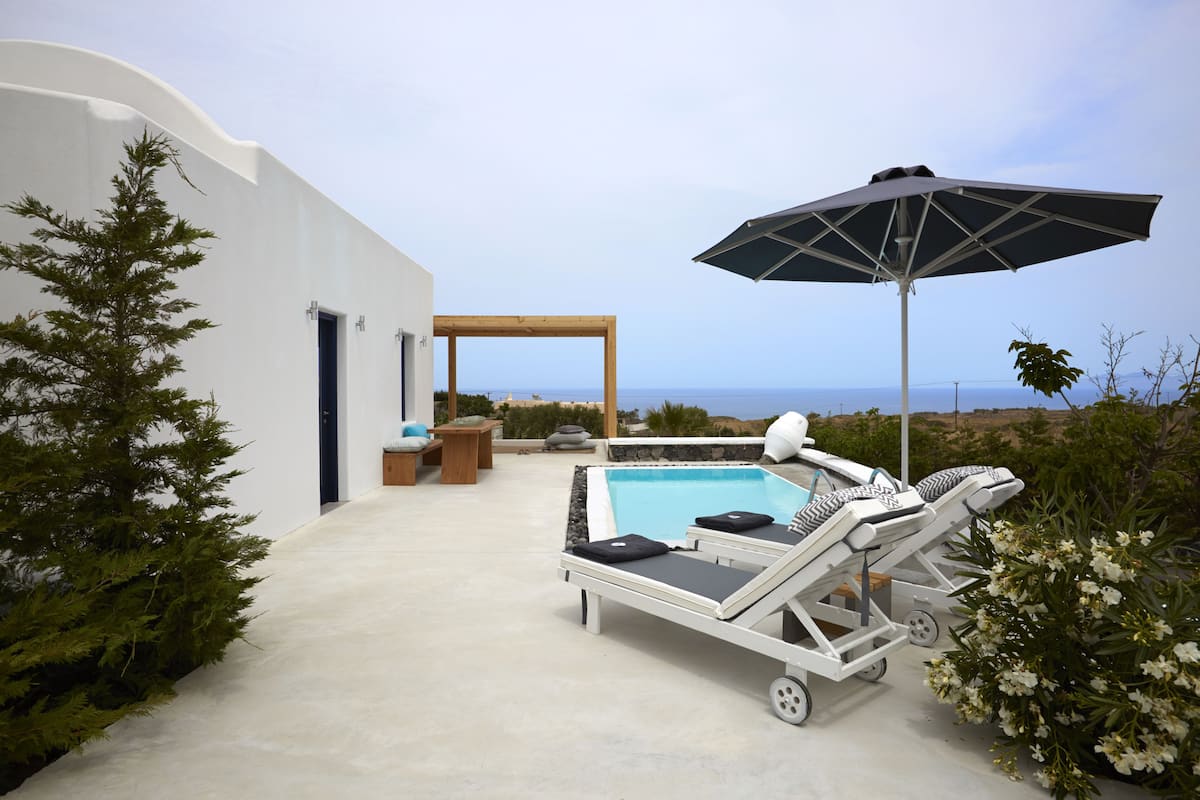 A modern outdoor pool area with lounge chairs and an umbrella, surrounded by a white building and natural landscape.