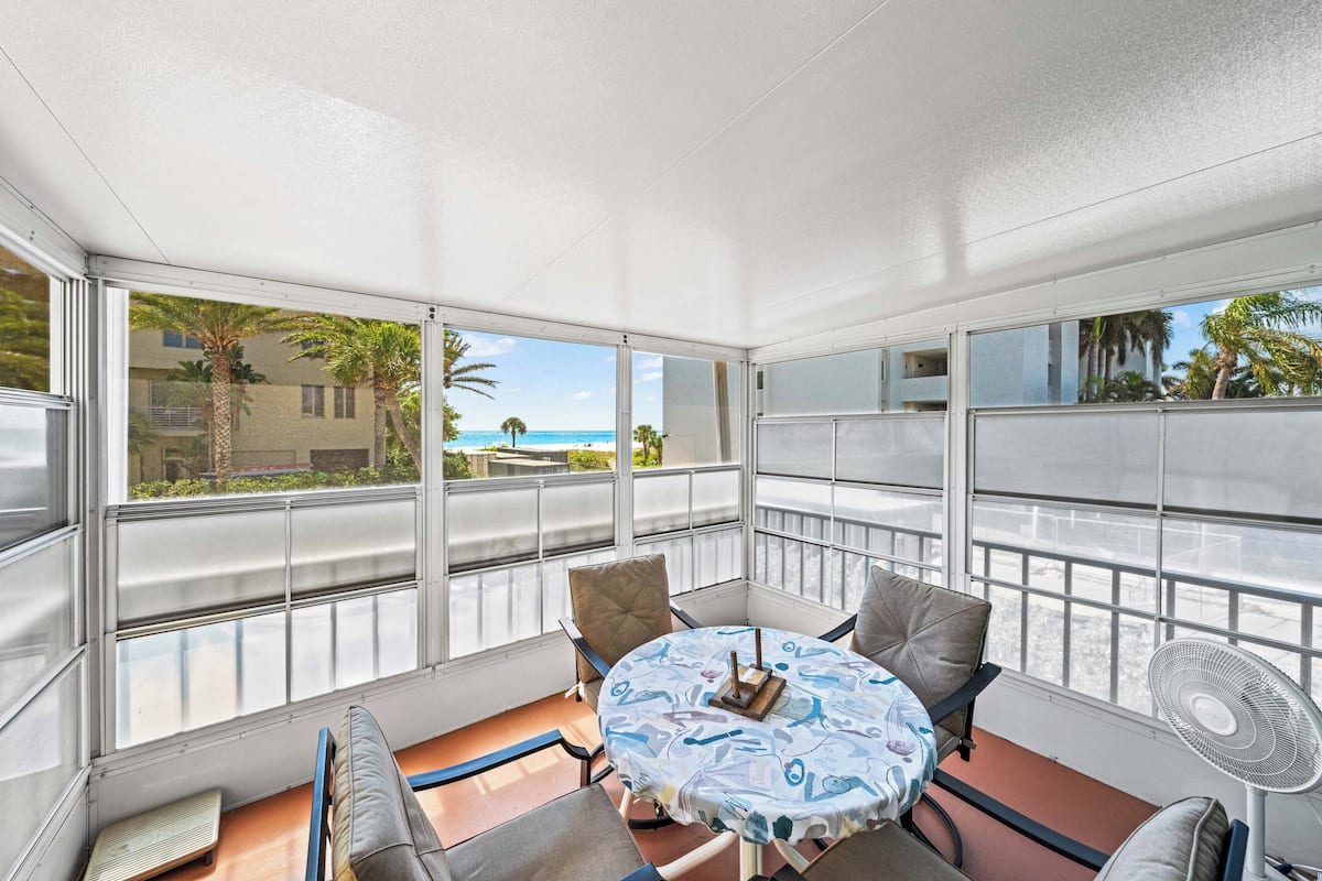 A sunroom with a table, chairs, a fan, and large windows overlooking a beach.