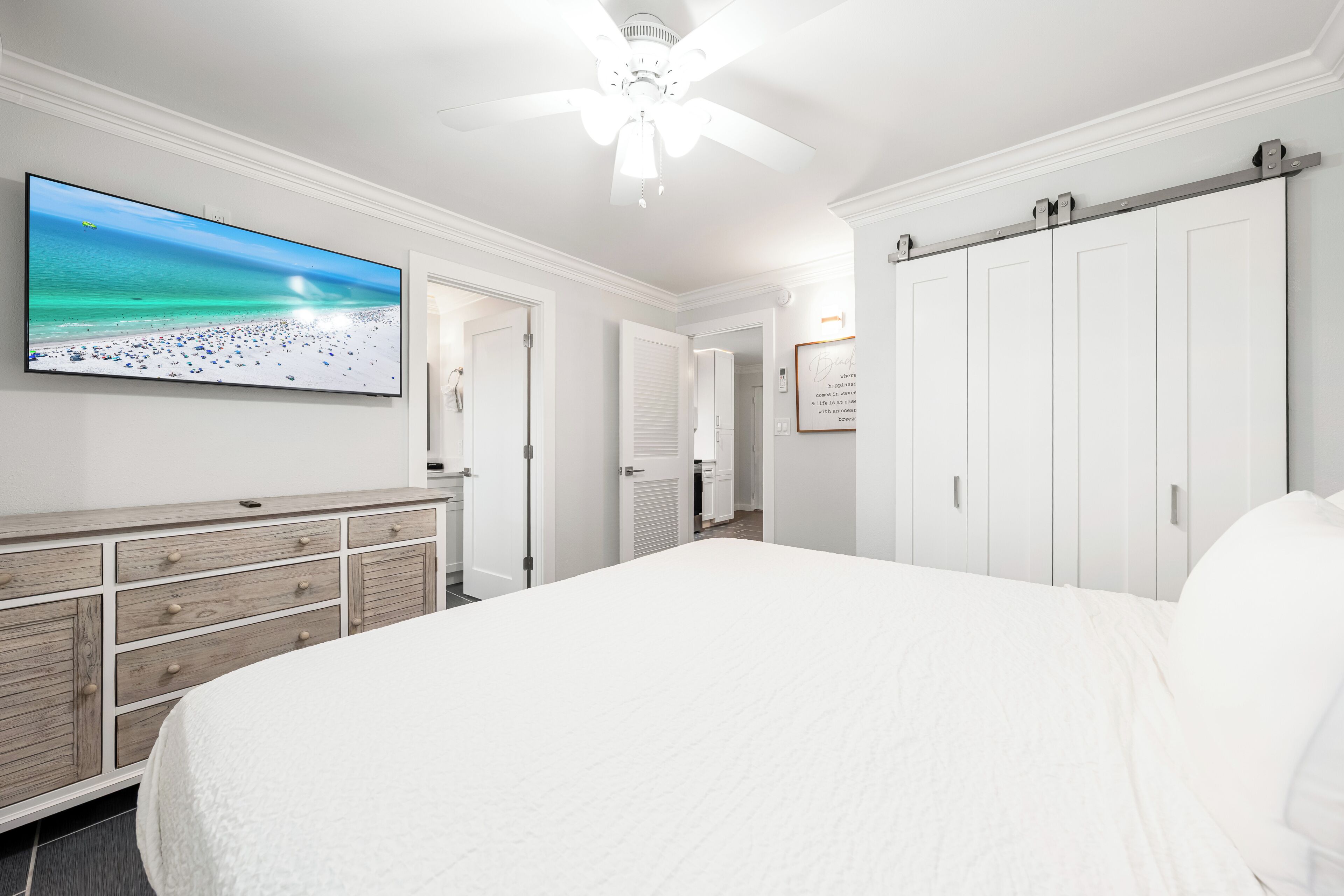 A bedroom with a large bed, a dresser, a TV on the wall, and a ceiling fan.
