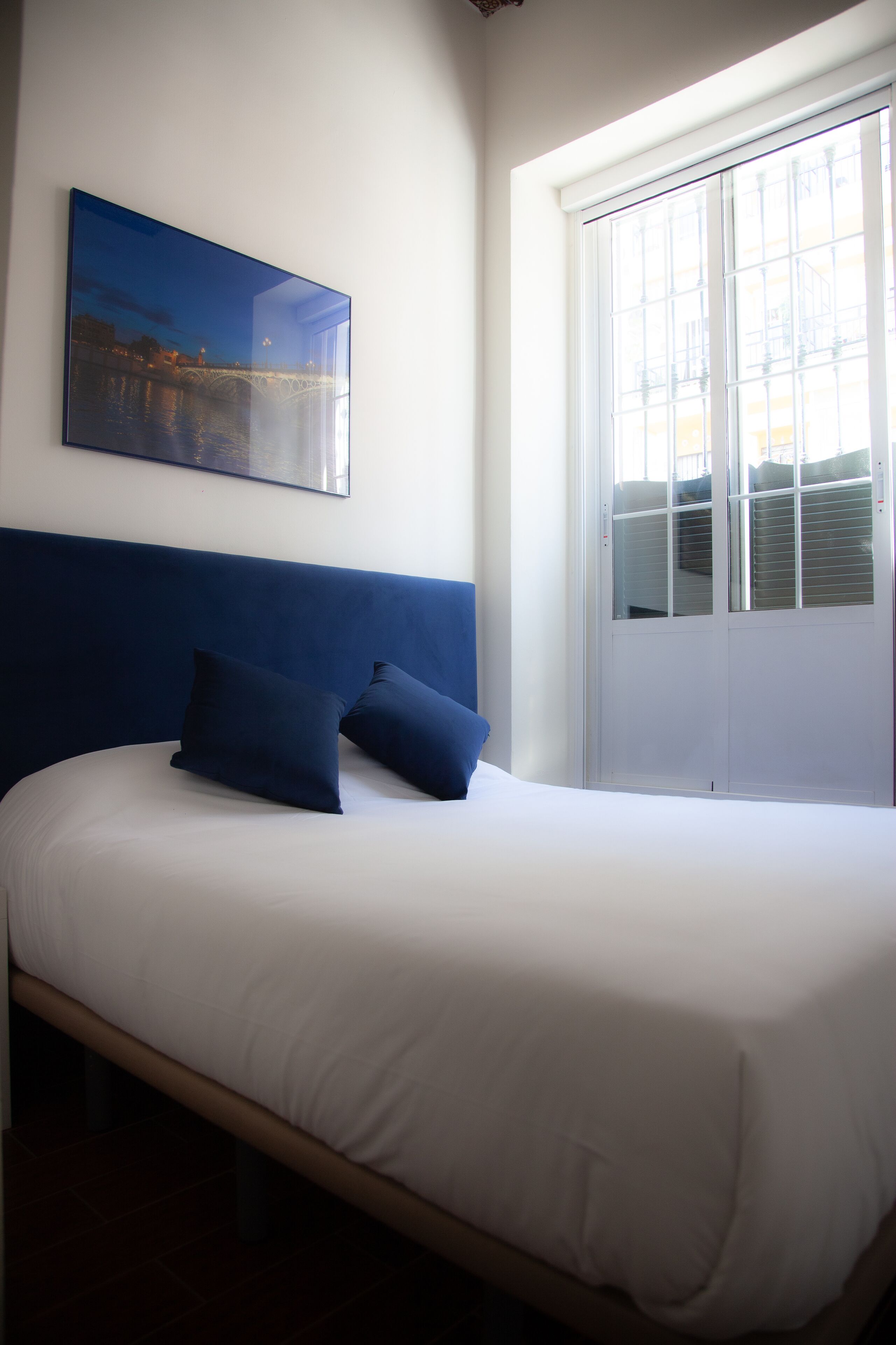 A bed with a white mattress and blue headboard, a window with blinds, and a painting of a bridge on the wall.