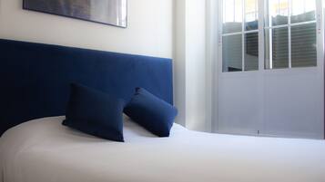 A bed with a white mattress and blue headboard, a window with blinds, and a painting of a bridge on the wall.