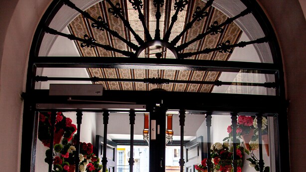 A wrought iron gate with floral decorations, leading to a tiled courtyard with a view of a street and buildings.