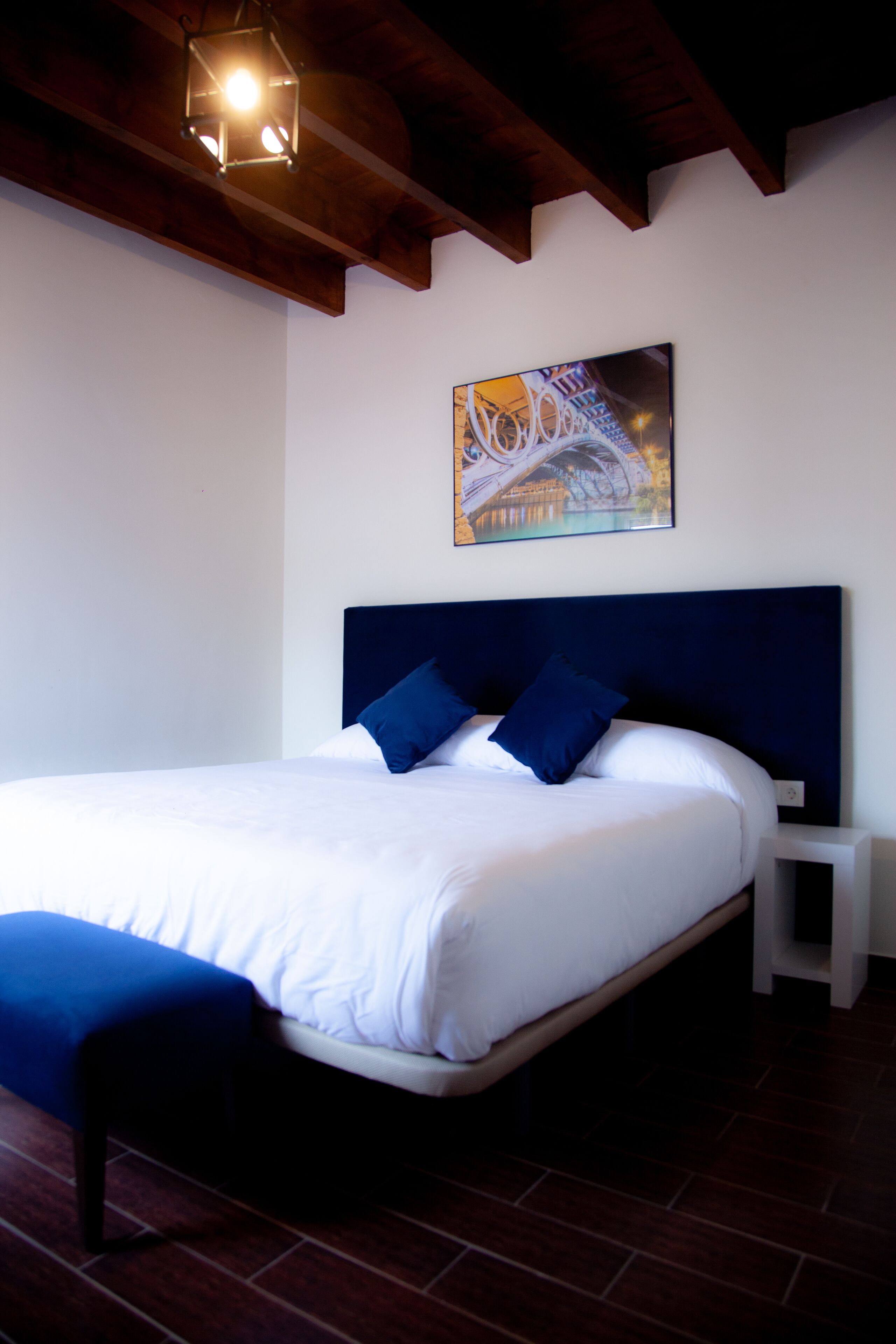 Una cama bien hecha con sábanas blancas y almohadas azules, una mesita de noche pequeña y un cuadro de un puente en la pared.