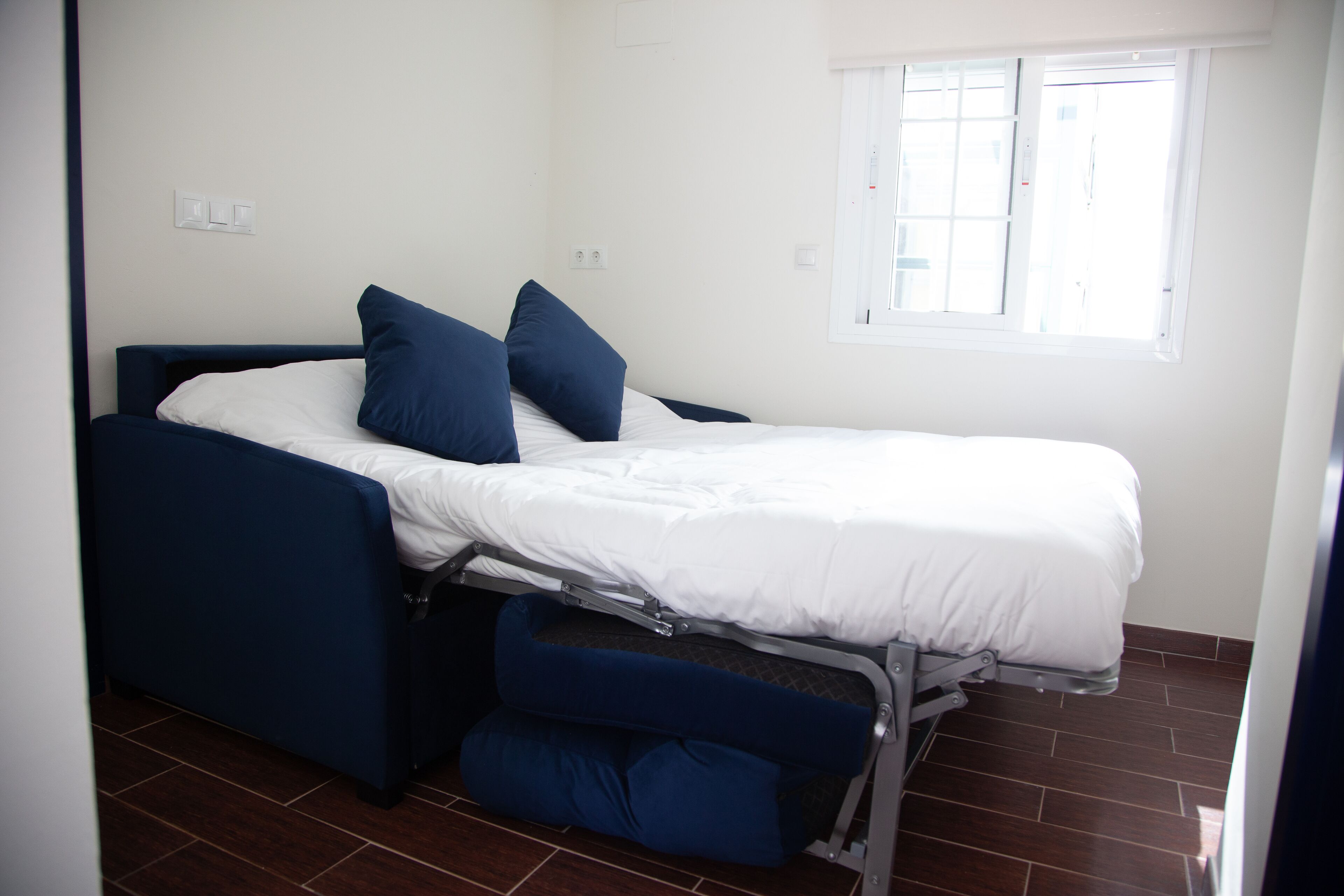 Una cama individual con colchón blanco y ropa de cama azul, un escritorio abatible y una ventana con persianas.