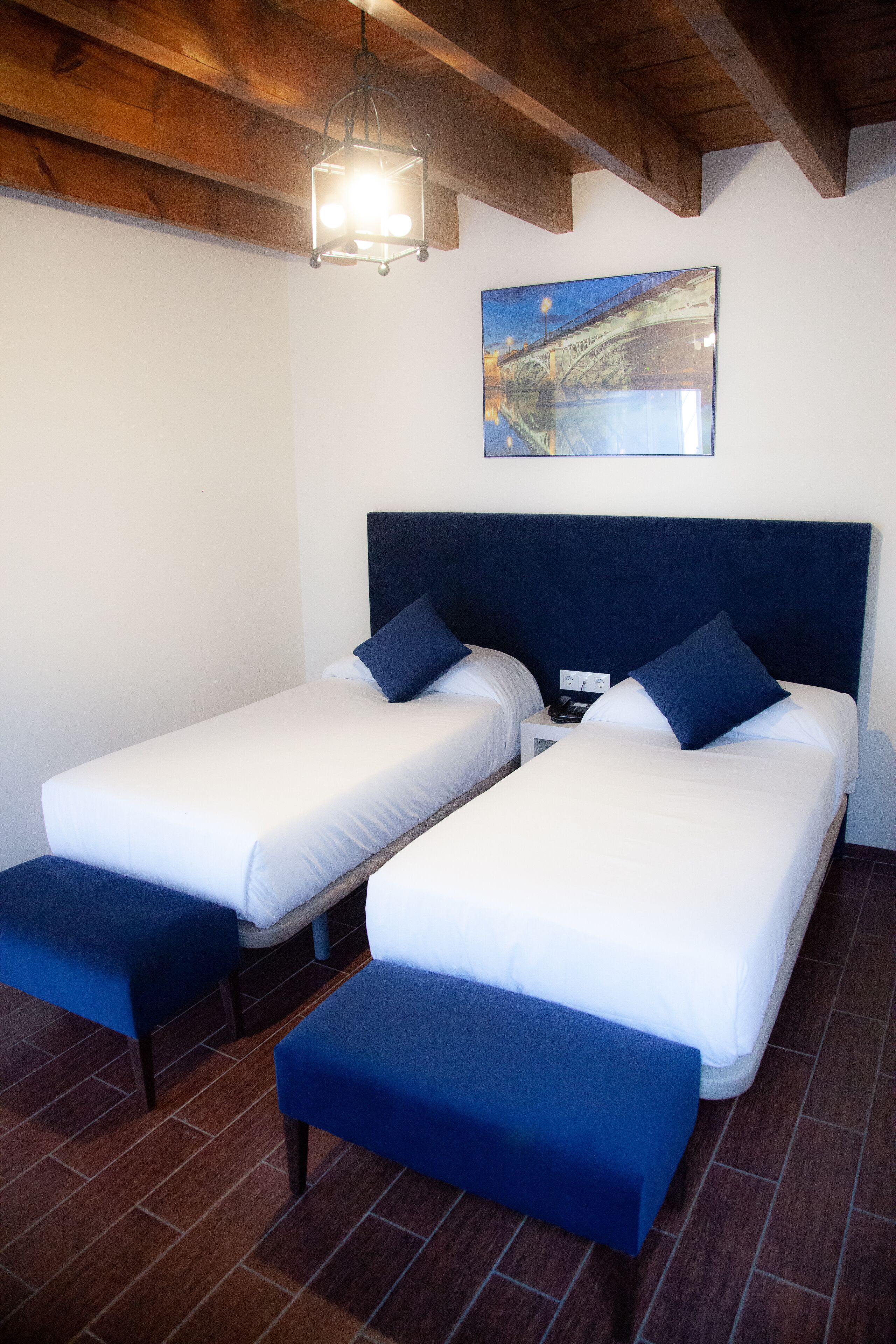 Habitación de hotel con dos camas individuales, cada una con cabecera azul y cojines a juego, techo de madera y un cuadro de un puente en la pared.