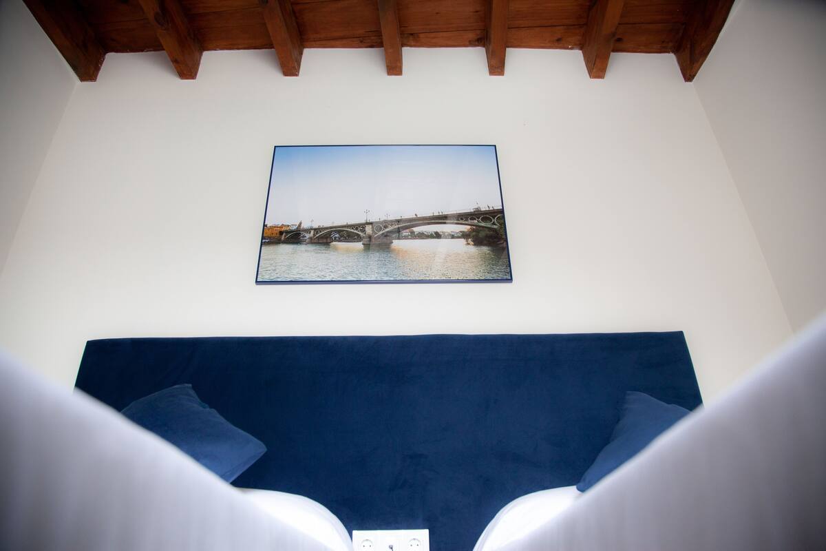 A room with a blue headboard, a framed picture of a bridge, and a white wall socket.