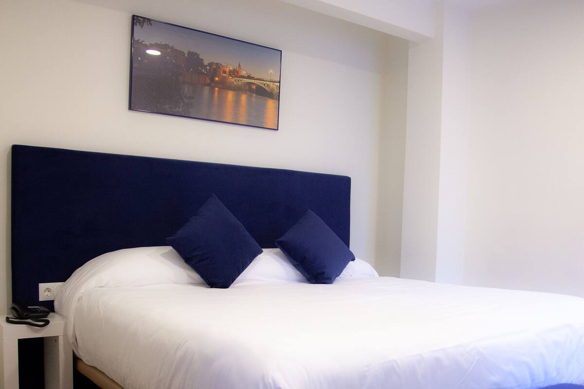 A neatly made bed with white linens and dark blue pillows, a bedside table with a phone, and a framed picture on the wall.