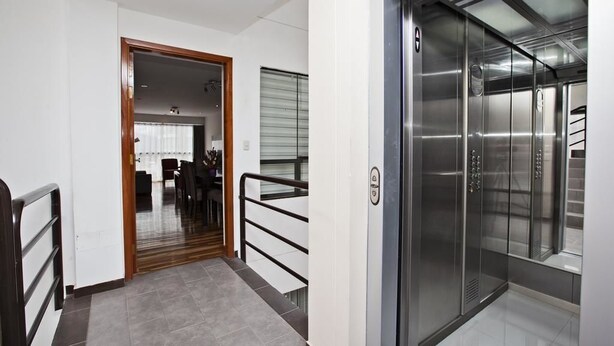 A modern elevator with metallic walls and a glass door, adjacent to a hallway with a wooden door and a dining area visible through a glass partition. - Polo Apartment (Cusco)