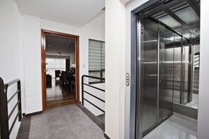 Un ascensor moderno con paredes metálicas y puerta de cristal, junto a un pasillo con puerta de madera y zona de comedor visible a través de un tabique de vidrio.