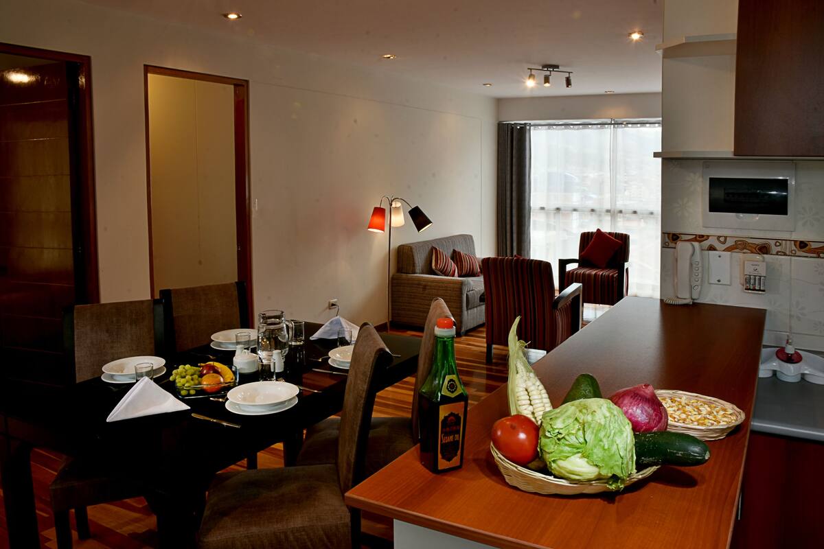 A dining area with a table set for two, a bottle of beer, and a basket of fresh vegetables.