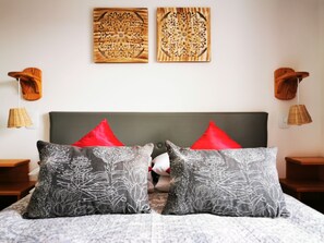 Ein Schlafzimmer mit grauem Kopfteil, zwei roten Kissen und zwei grauen Kissen mit floralem Muster. Oberhalb des Kopfteils befinden sich zwei Holz-Wandbilder.