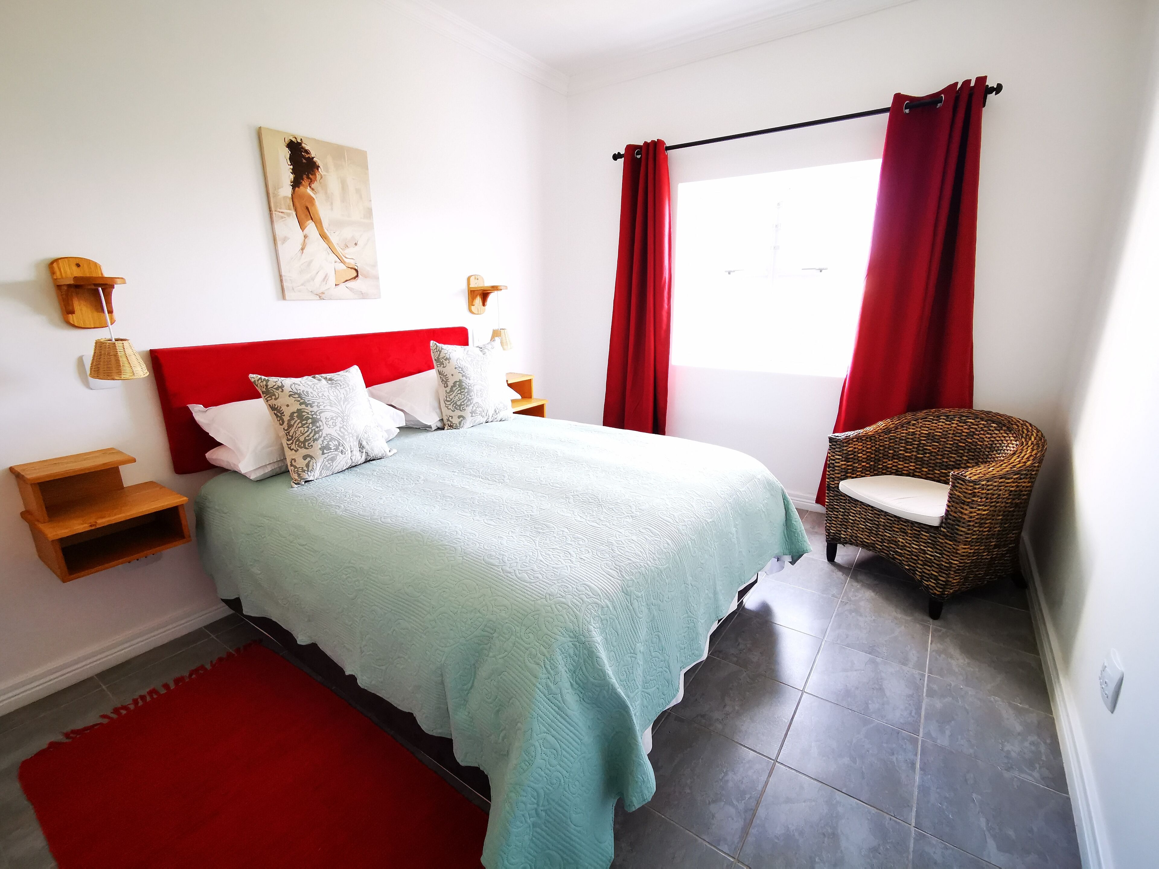 A bedroom with a bed, a red headboard, a wicker chair, a red rug, and a painting on the wall.