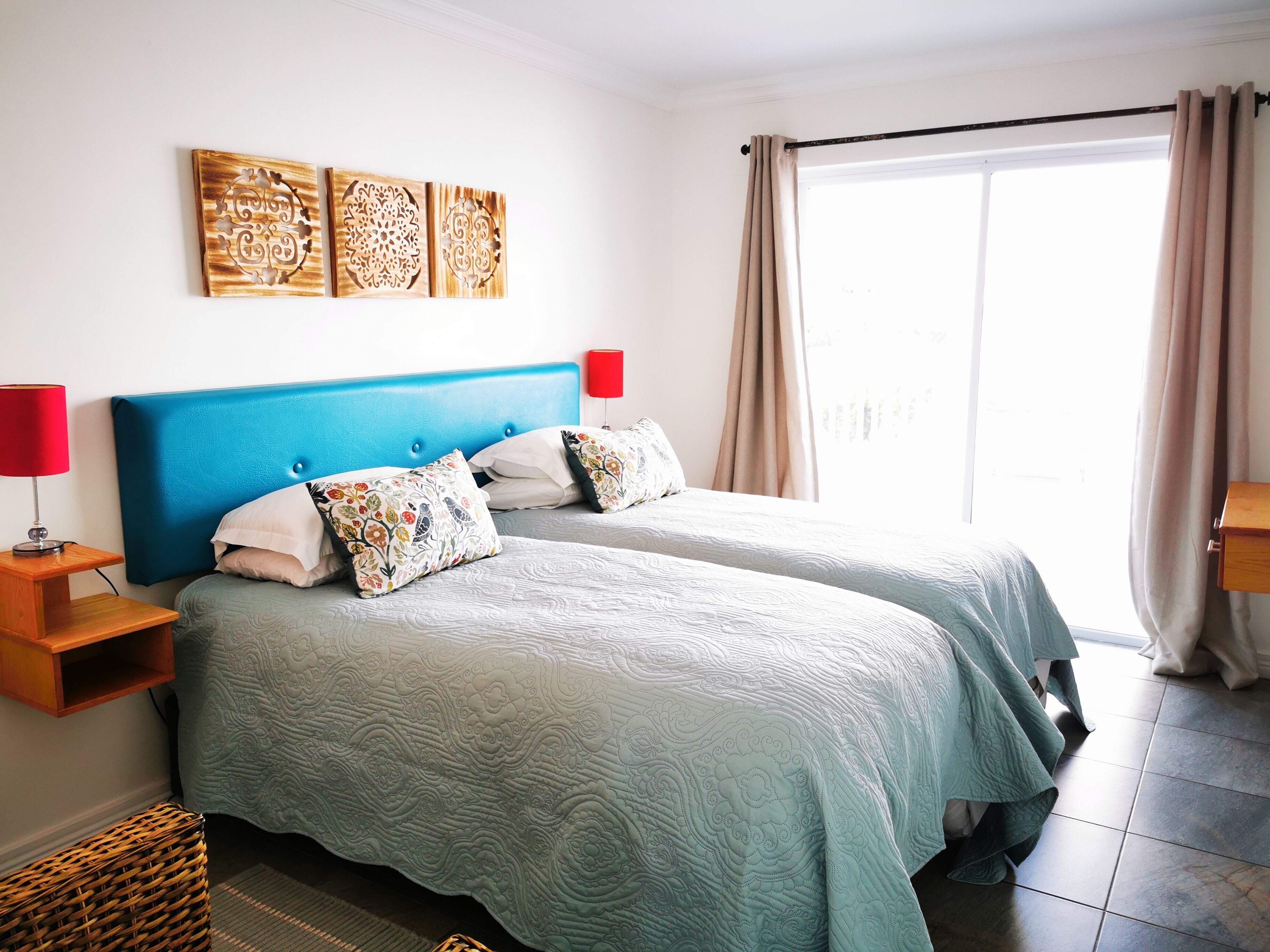 A bedroom with two beds, a blue headboard, a red lamp, and a large window with curtains.