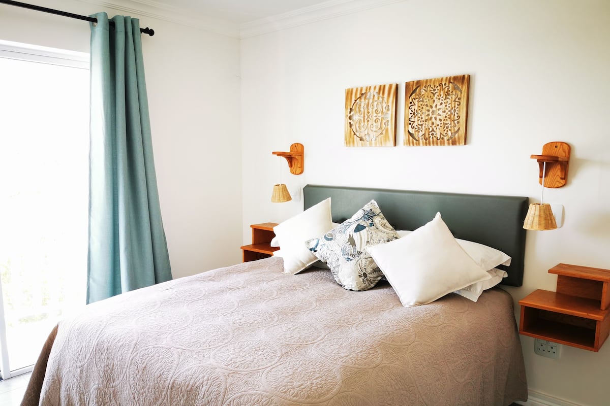 Ein ordentlich eingerichtetes Schlafzimmer mit Bett, Kissen, Kopfteil, Wandbildern und Nachttischlampen.