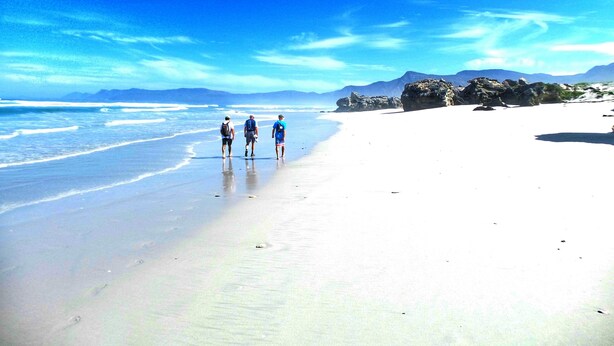 Een strand met drie mensen die langs de kust lopen, een heldere lucht en verre bergen.