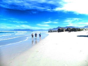 Une plage où trois personnes se promènent le long du rivage, sous un ciel dégagé, avec des montagnes qui se dessinent à l’horizon.
