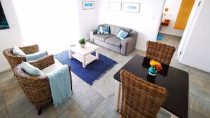 Een woonkamer met een grijze bank, rieten stoelen, een salontafel en een eettafel met een vaas.