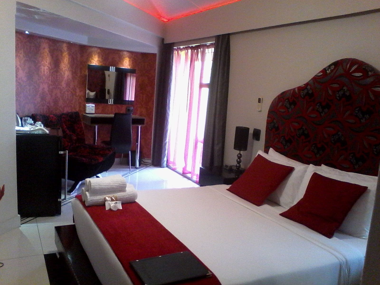 A modern hotel room with a large bed, red and white bedding, a desk with a chair, a mirror, and a window with curtains.