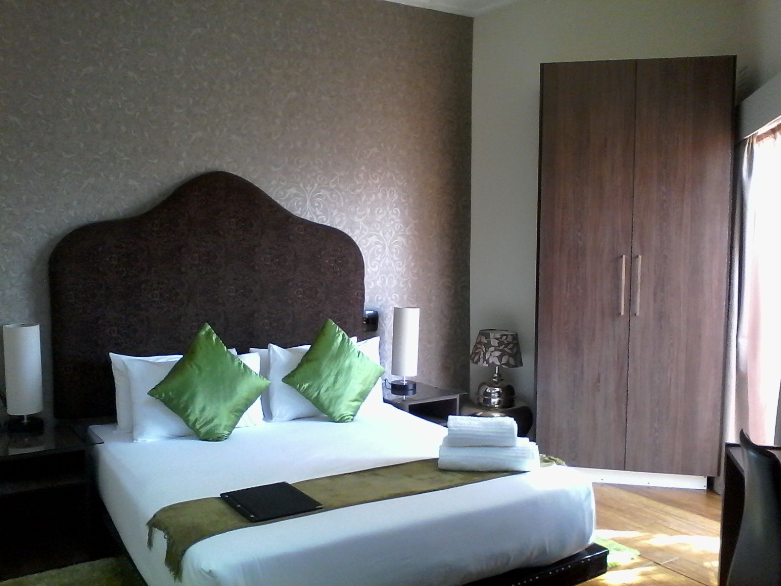 A hotel room with a large bed, two nightstands, a lamp, a wardrobe, and a view of the outside.