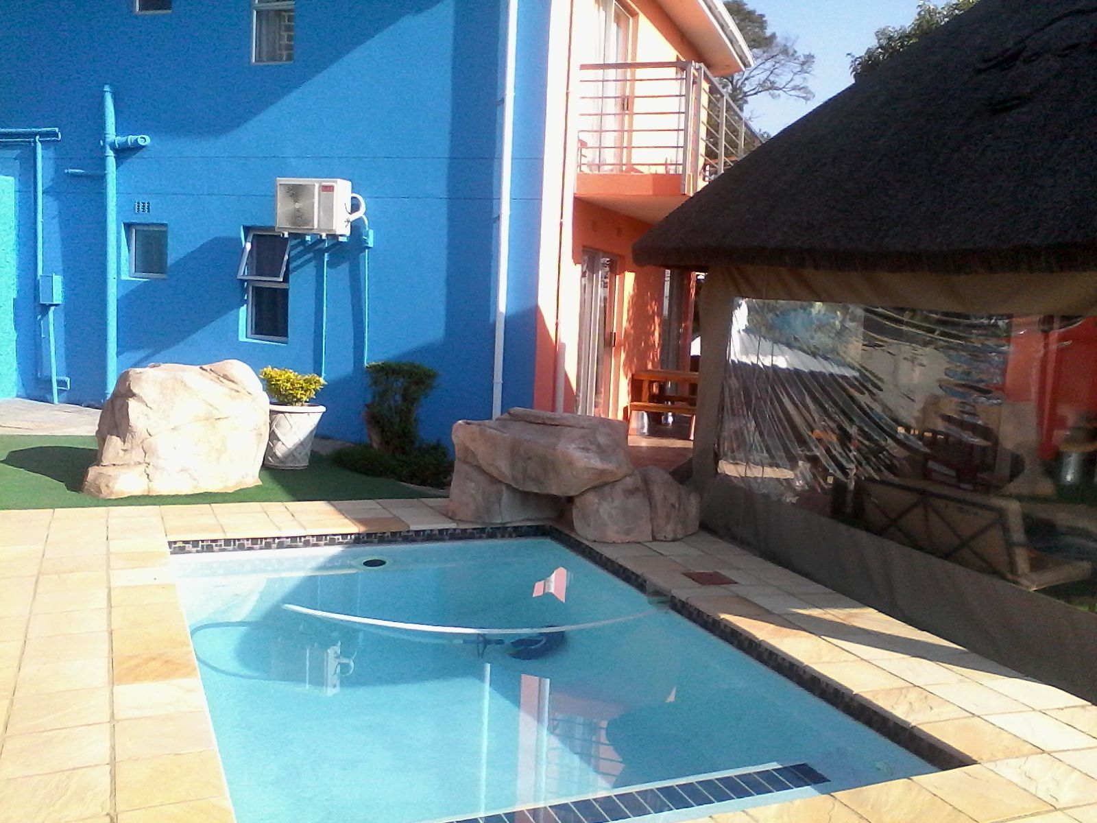 Una piscina con solárium de baldosas, rocas grandes y un edificio con paredes azules y un balcón rojo.