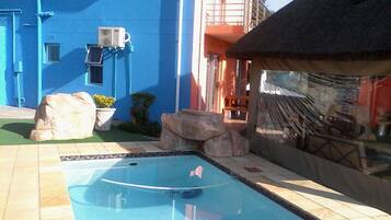Una piscina con solárium de baldosas, rocas grandes y un edificio con paredes azules y un balcón rojo.