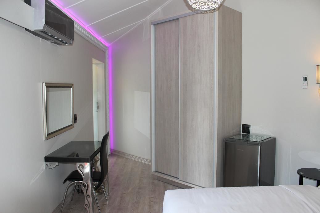 A modern hotel room with a bed, a desk with a chair, a wardrobe, a small bathroom with a mirror, and a ceiling light fixture.