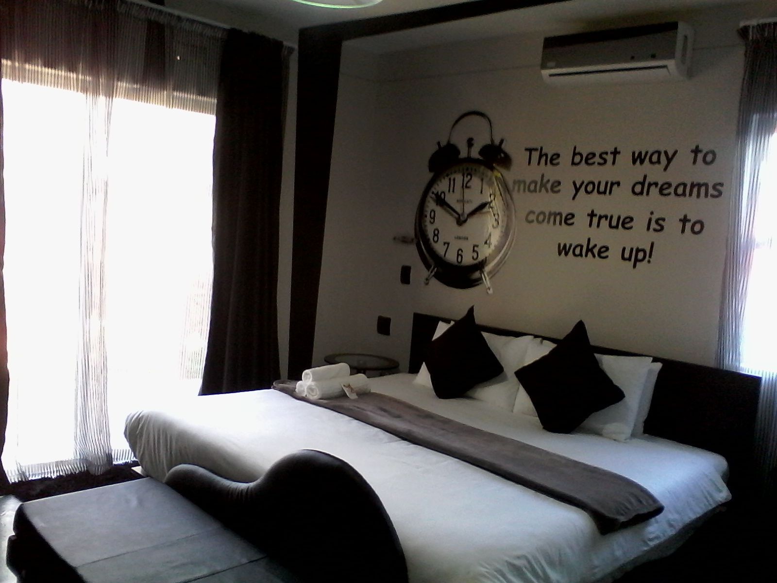 A hotel room with a bed, a clock, and a motivational quote on the wall.