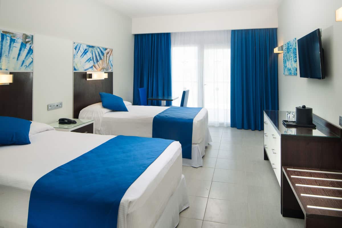 A hotel room with two beds, a desk, a chair, a TV, and artwork on the walls.
