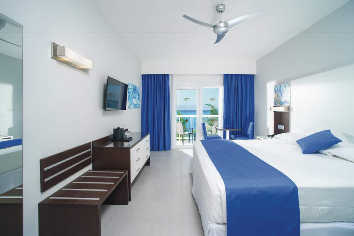 A hotel room with a bed, a desk, a chair, a television, and a balcony with a view of the ocean.