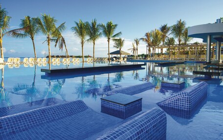 A pool with tiled lounge areas, palm trees, and a clear sky.