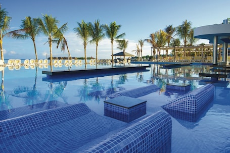 A pool with tiled lounge areas, palm trees, and a clear sky.