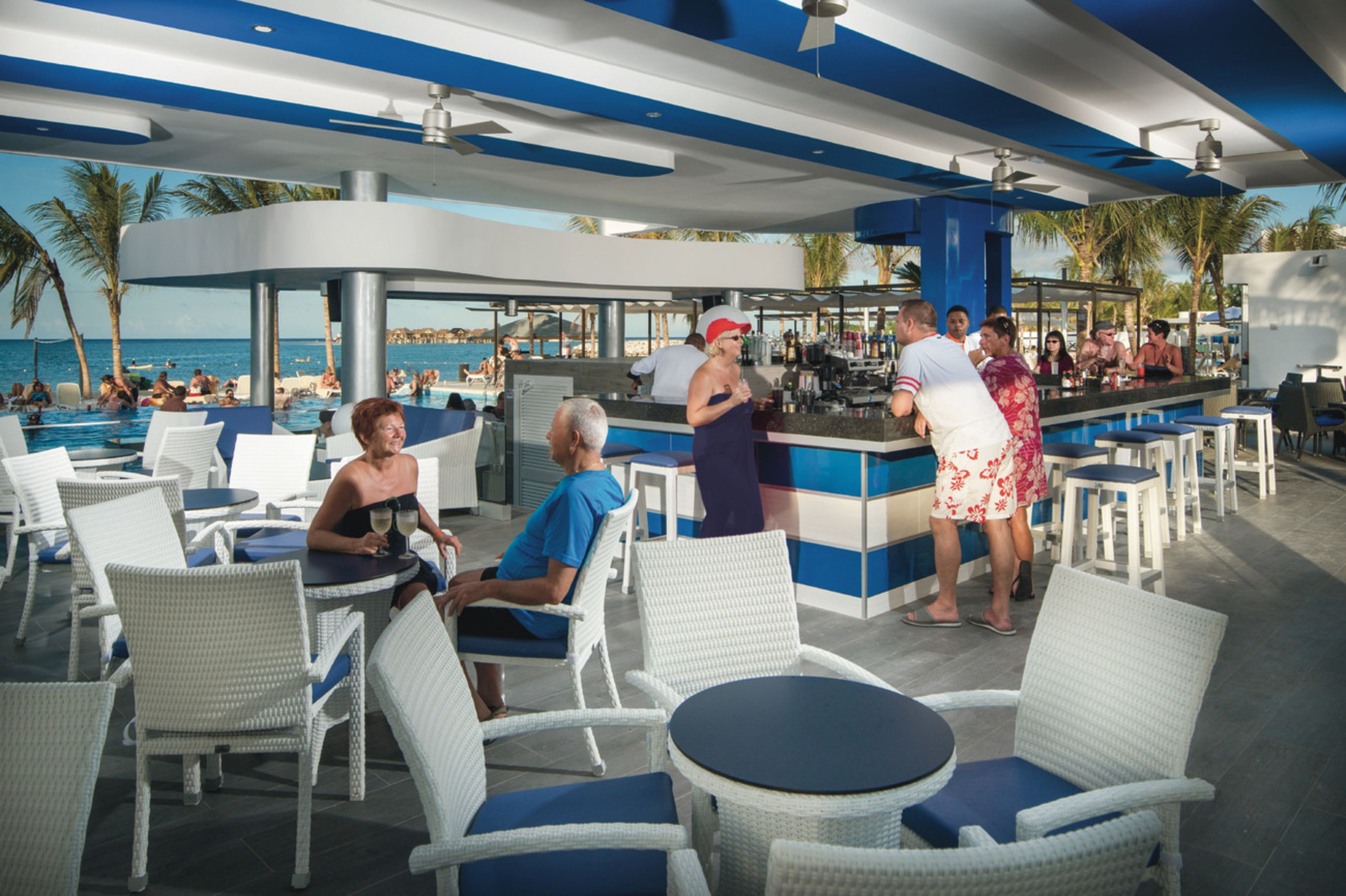 A poolside bar area with white wicker chairs and tables, a bar counter, and a view of the ocean.