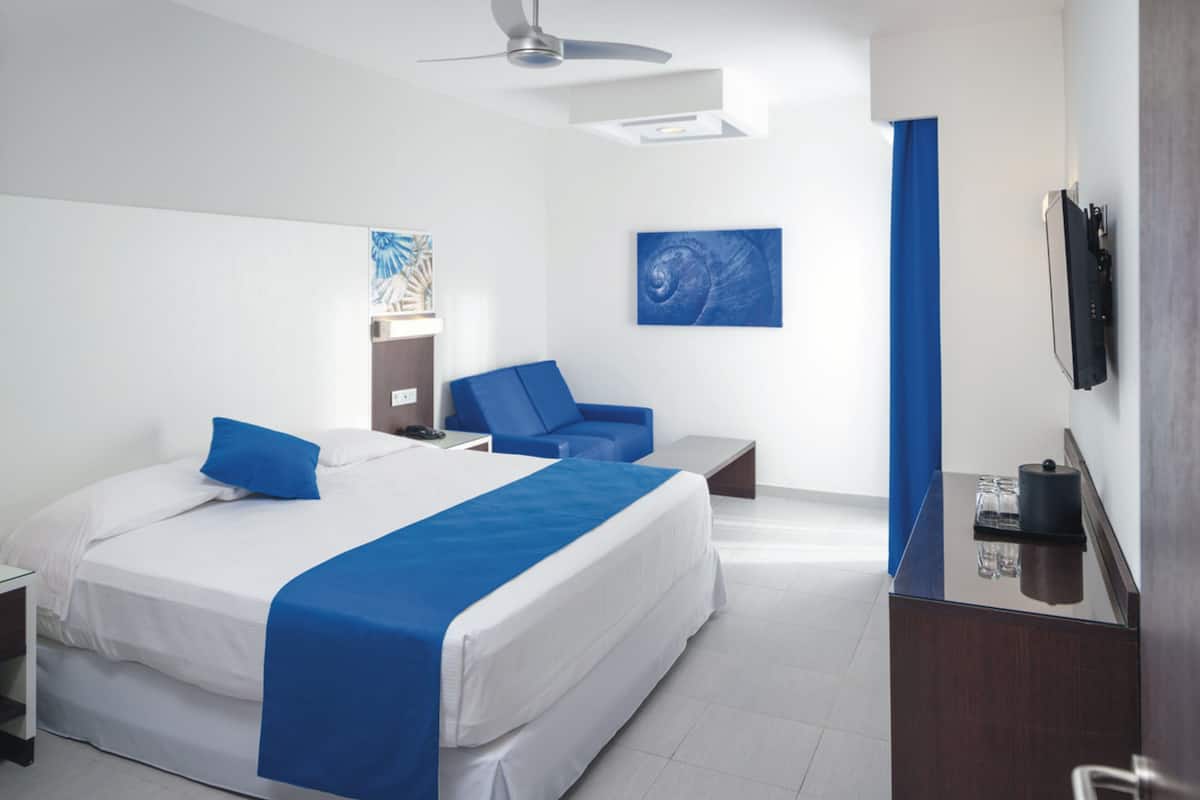 A modern hotel room with a large bed, a desk, a chair, a ceiling fan, and artwork on the walls.
