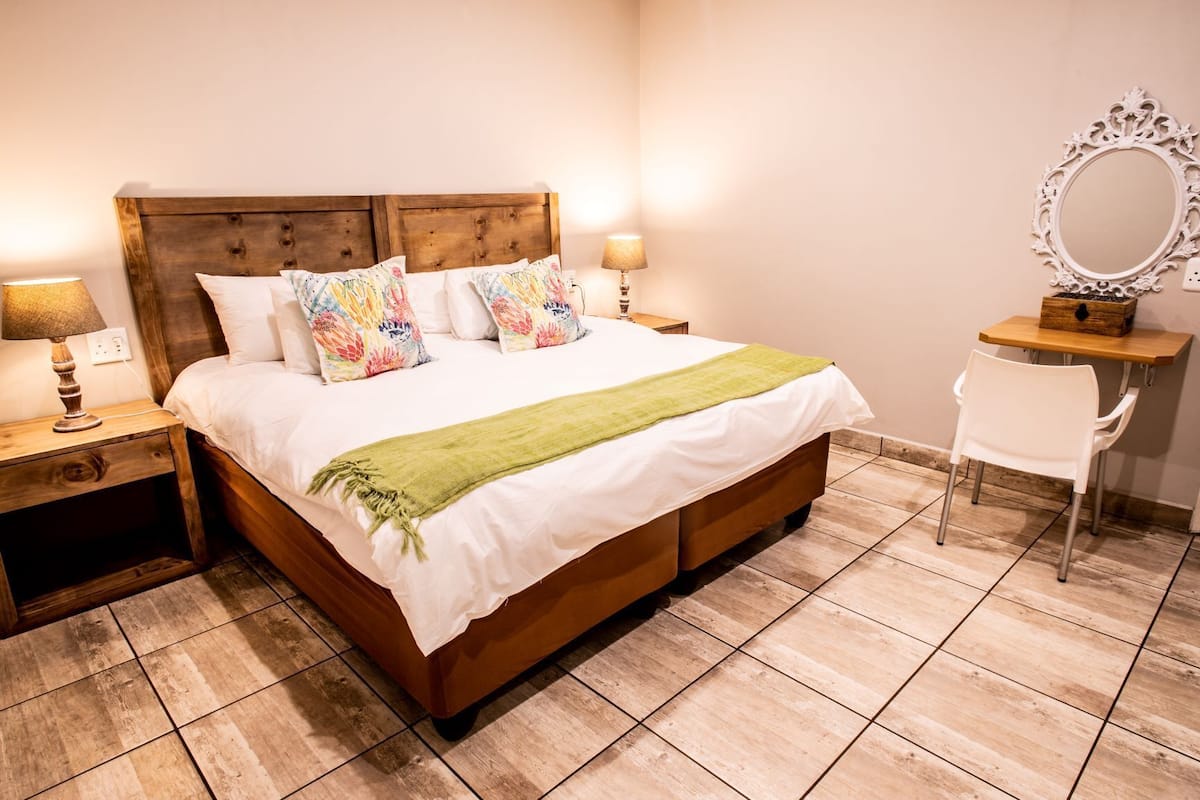 Quarto com cama de madeira, roupa de cama branca, manta verde, duas luminárias de cabeceira, uma cadeira branca e um espelho sobre uma mesa de madeira.