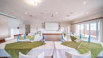 Ein Konferenzraum mit weißen Tischen, grünen Tischdecken und weißen Stühlen. Auf den Tischen stehen Wasserflaschen und im Hintergrund ist ein Projektionsschirm zu sehen.