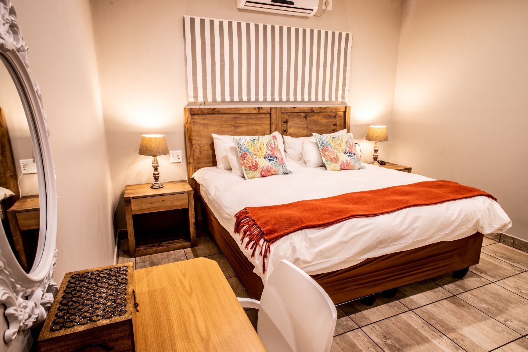 A neatly arranged bedroom with a wooden bed, two bedside tables with lamps, a mirror, and a small wooden table with a decorative item.