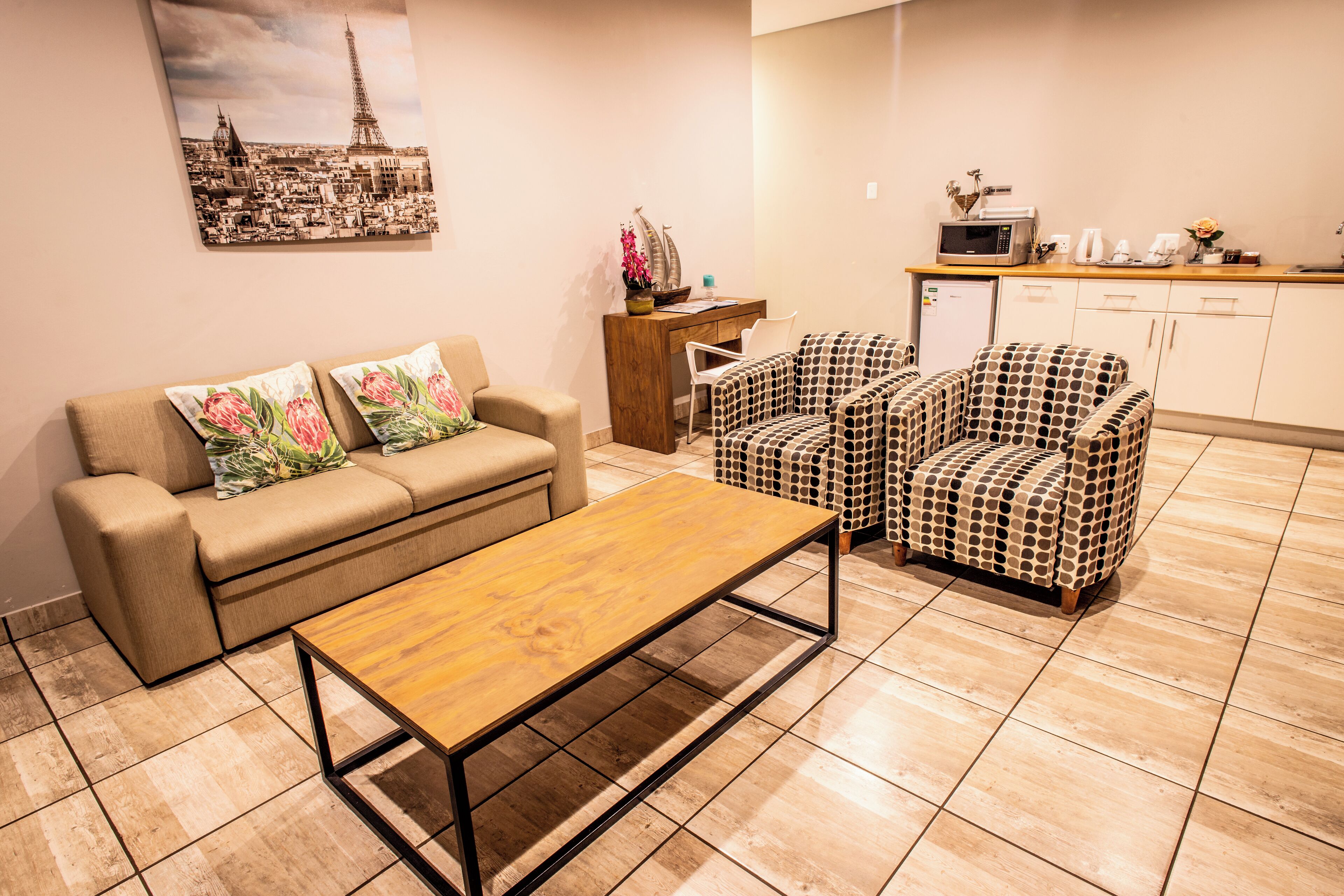 Een woonkamer met een bank, twee fauteuils met een patroon, een salontafel, een eethoek met een houten tafel, en een keuken met een magnetron en kastjes.