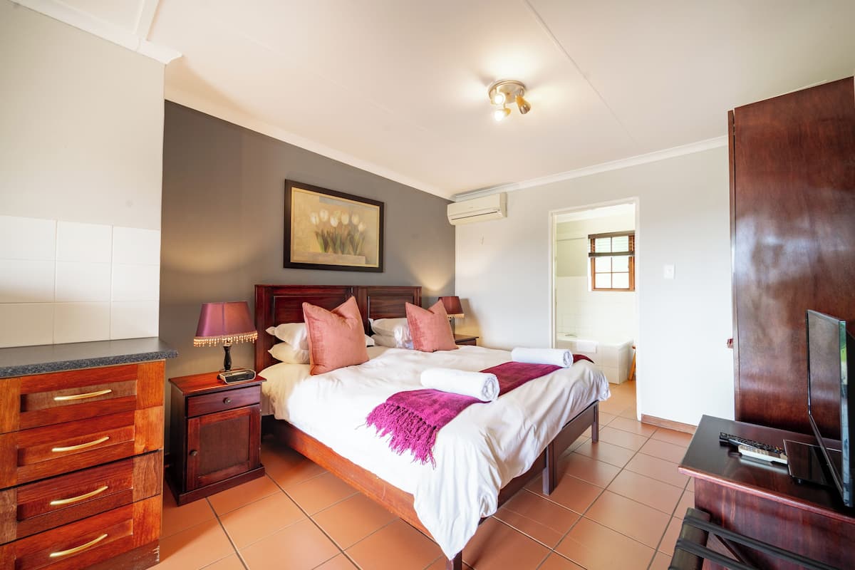 Quarto de hotel com cama, mesas de cabeceira de madeira, escrivaninha com televisão e um quadro na parede.
