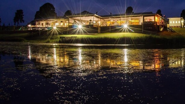 A large, illuminated building with a reflective water surface in front, surrounded by trees. - St Ives Lodge (Lidgetton West)