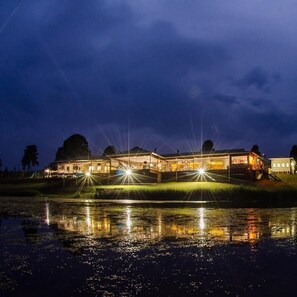 A large, illuminated building with a reflective water surface in front, surrounded by trees. - St Ives Lodge (Lidgetton West)