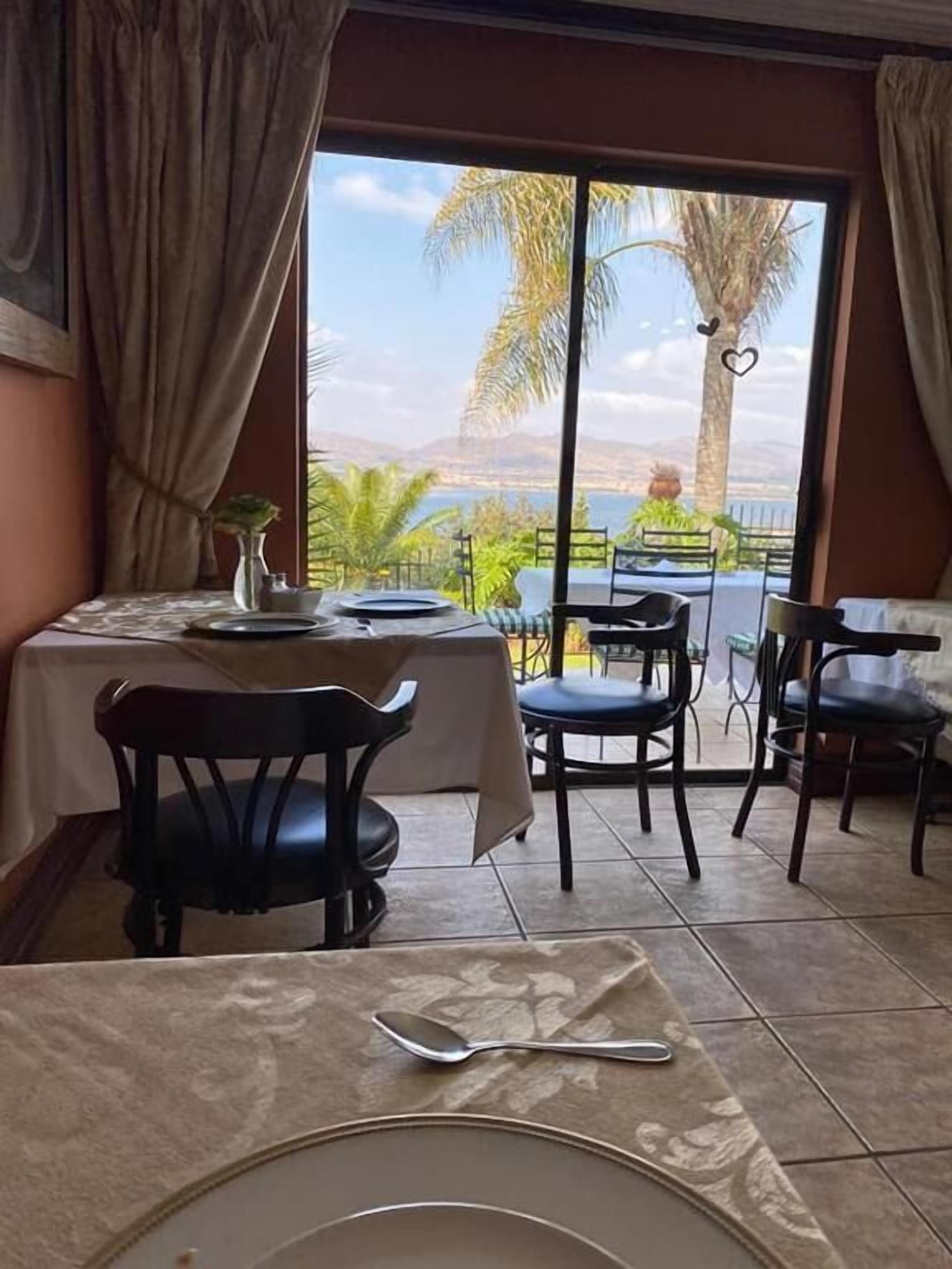 A dining area with a table set for two, a view of palm trees and mountains through large windows, and a balcony with chairs.
