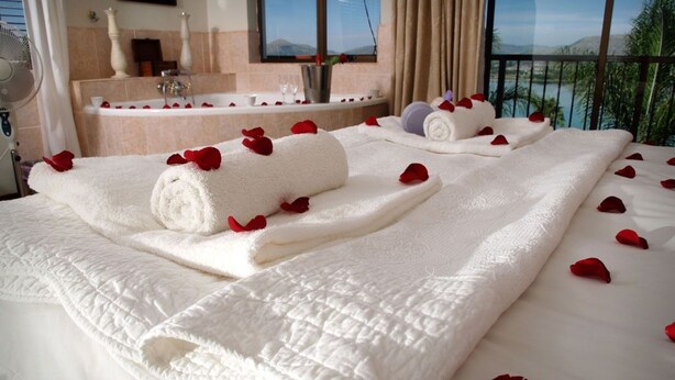 A hotel room with a bed adorned with white towels and rose petals, a bathtub, and a view of the sea. - La Dolce Vita Guest House (Hartbeespoort)