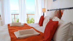A hotel room with a bed, white linens, an orange throw blanket, red pillows, and a view of the ocean. - La Dolce Vita Guest House (Hartbeespoort)