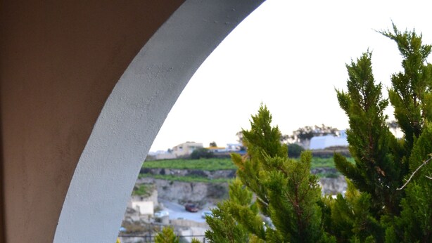 A balcony with a table and two glasses of red wine, overlooking a green landscape and buildings. - Floralia (Santorini)